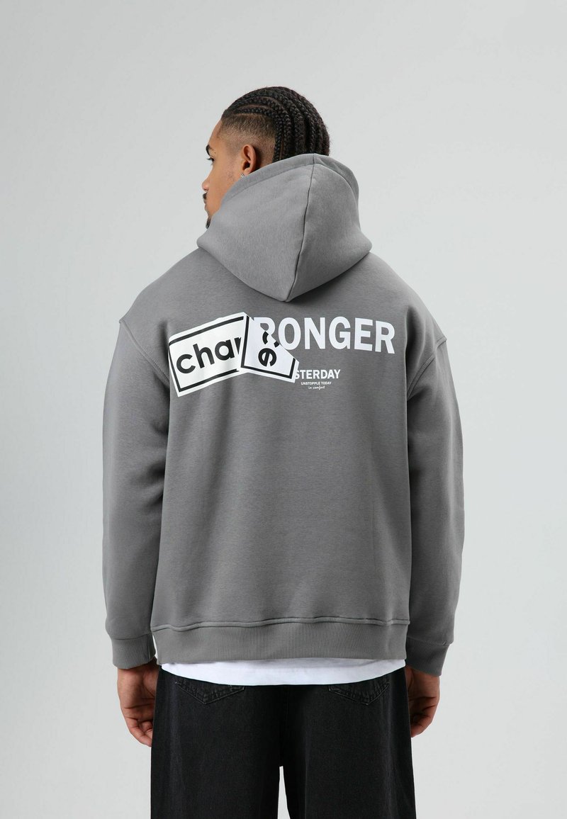 Grijze hoodie met losse pasvorm, met gedurfde witte tekst op de achterkant. Zachte textuur en geribbelde boorden maken het ontwerp compleet.