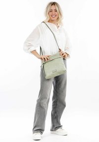 Borsa a tracolla in pelle verde chiaro con pattina, hardware argentato e tracolla regolabile, abbinata a una camicia bianca e jeans grigi.