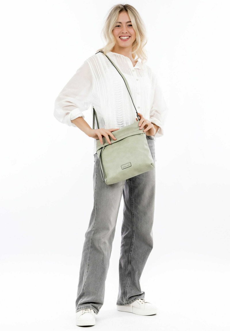 Borsa a tracolla in pelle verde chiaro con pattina, hardware argentato e tracolla regolabile, abbinata a una camicia bianca e jeans grigi.