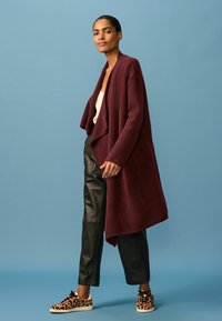 Burgunderfarbener oversized Cardigan aus weichem Material, über einem cremefarbenen Top getragen. Kombiniert mit schwarzen Lederhosen und gemusterten Turnschuhen.