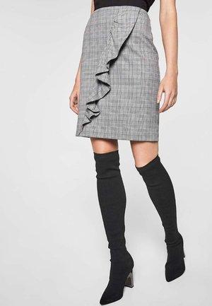 Femme portant une jupe grise à carreaux longueur genou avec un détail volanté à l'avant et des bottes noires à talons au-dessus du genou sur fond uni.