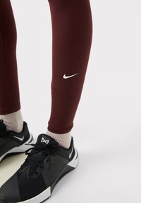 Burgundi leggings fehér Nike logóval, fekete, részletes textúrájú és fehér kiegészítőkkel ellátott sportcipővel párosítva. A zoknik világos bézs színűek.
