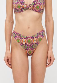 Femme portant un maillot de bain violet avec un haut et un bas ornés de motifs floraux et géométriques brillants en orange, vert et rouge, vue des épaules jusqu'aux cuisses supérieures.
