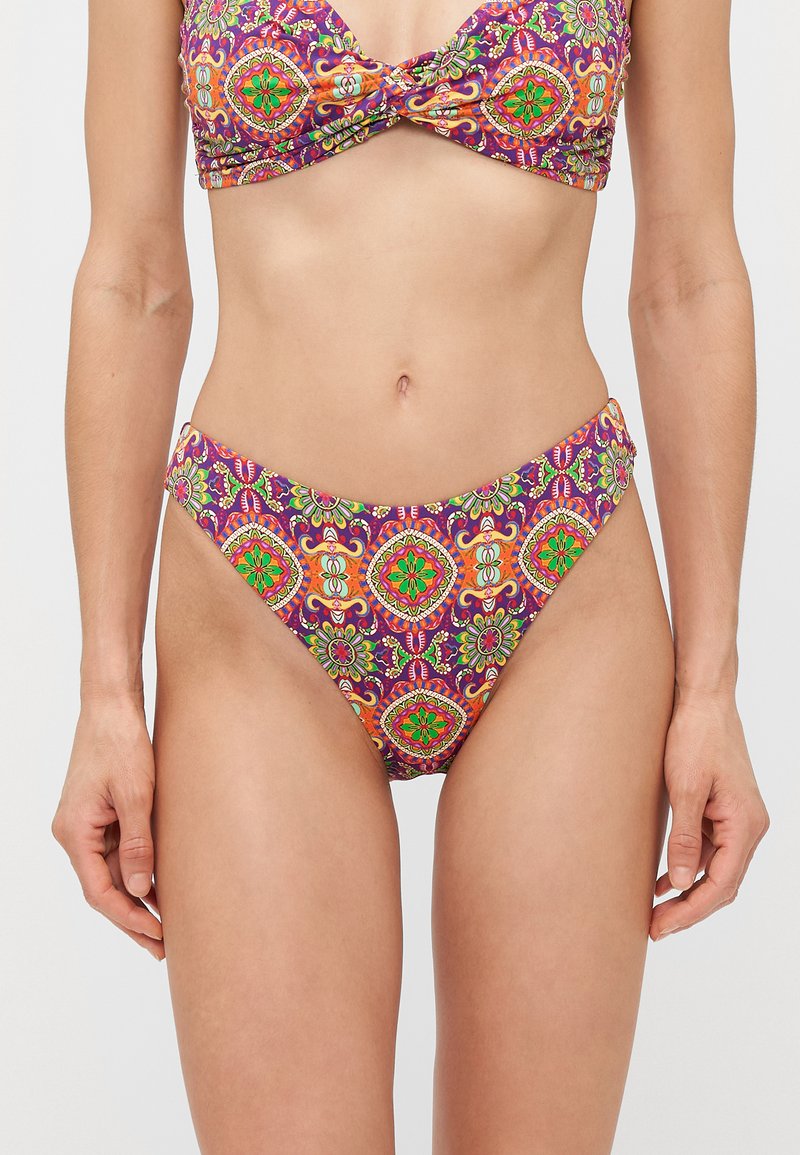 Femme portant un maillot de bain violet avec un haut et un bas ornés de motifs floraux et géométriques brillants en orange, vert et rouge, vue des épaules jusqu'aux cuisses supérieures.