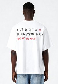 Balts pārāk liels T-krekls ar melnu un sarkanu tekstu aizmugurē: "MAZLIET ♥ ŠAJĀ BRUTĀLAJĀ PASAULĒ (BET NE PĀRSPĒJOT)."