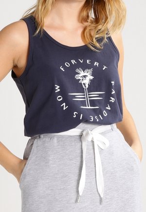 Kobieta nosząca granatowy top bez rękawów z białą grafiką palmy i okrągłym tekstem, w połączeniu z jasnoszarymi spodniami ze sznurkiem.