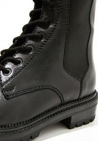 Černá kotníková bota z kůže s texturovaným látkovým panelem, výrazným šněrováním a pevným podrážkovým vzorem. Má viditelné švy a robustní design.