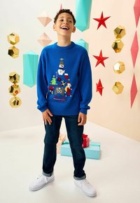 Pulover tricotat de culoare albastră, cu un design tematic de sărbători, prezentând un om de zăpadă, copaci și animale. Asortat cu blugi închiși la culoare și adidași albi.