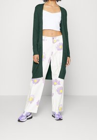 Vihreä neulottu pitkä neuletakki, valkoiset housut, joissa on violettia ja keltaista kukka-ommelia, sekä violetteja tennareita korostavat rentoa ilmettä.