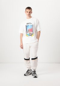 Camiseta blanca de gran tamaño con un diseño gráfico colorido que presenta un paisaje costero. Combinada con pantalones beige claros con rayas negras y beige.