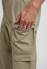 Beige Outdoor-Cargo-Hosen bestehen aus einem strukturierten Stoff, haben seitliche Taschen und einen lockeren Schnitt, wobei eine Hand in der offenen Tasche liegt.