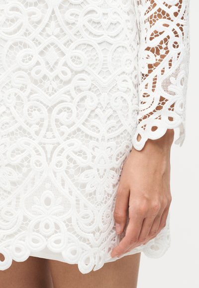 Gros plan d’une main reposant sur une robe en dentelle blanche avec des motifs floraux et géométriques complexes sur la manche et l’ourlet.