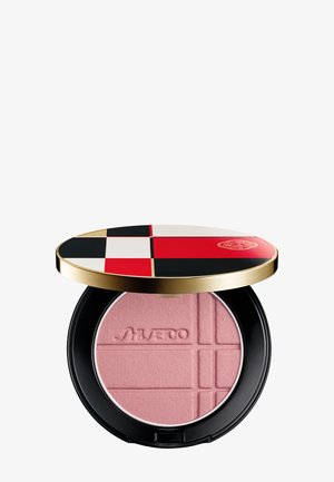 Poudre compacte avec teinte rose blush, embossée du nom « Shiseido », dans un boîtier noir et or avec un couvercle à motif géométrique rouge, noir et blanc.