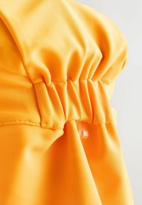 Élénk narancssárga szövet összegyűrt mintával és varrott részletekkel. Látható gombbal és sima textúrával rendelkezik.