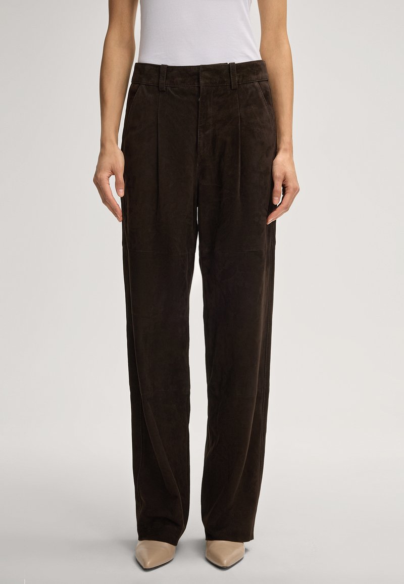 Pantalons larges en daim marron avec une texture lisse, des plis devant et une taille haute. Portés avec des chaussures beiges à bout pointu pour le contraste.