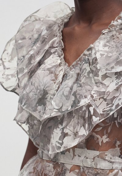 Διαφανές γκρι φλοράλ μπλούζα με βολάν, στρωμένο ύφασμα και V-λαιμόκοψη, που φοριέται από άτομο με σκουρόχρωμο δέρμα.