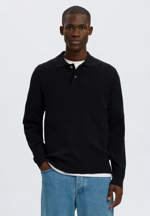 Homme portant un pull polo noir côtelé à manches longues sur une chemise blanche avec un jean bleu, debout devant un fond clair uni.