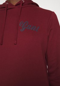 Sudadera burdeos con cordones, con un logo azul "Gant" ligeramente elevado en el pecho. Textura de tela suave.