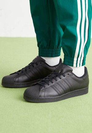 Personne portant des baskets noires à bout renforcé et un pantalon de survêtement vert avec des bandes blanches sur un sol vert.