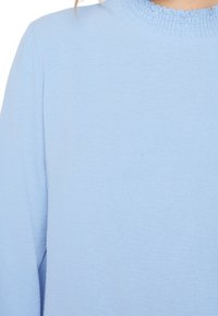 Top azzurro chiaro a maniche lunghe con scollo texturizzato e arricciato. Tessuto liscio con un aspetto morbido e arioso e una lucentezza sottile.