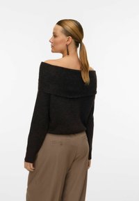 Off-shoulder, mörkstickad tröja med en tjock krage, kombinerad med ljusbruna vida byxor som har bakfickor och knappar.