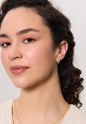 Złote kolczyki w kształcie obrączek o zaokrąglonym designie, z małym zdobieniem. Gładka tekstura; noszone w przekutym uchu. Ton skóry wydaje się naturalny.