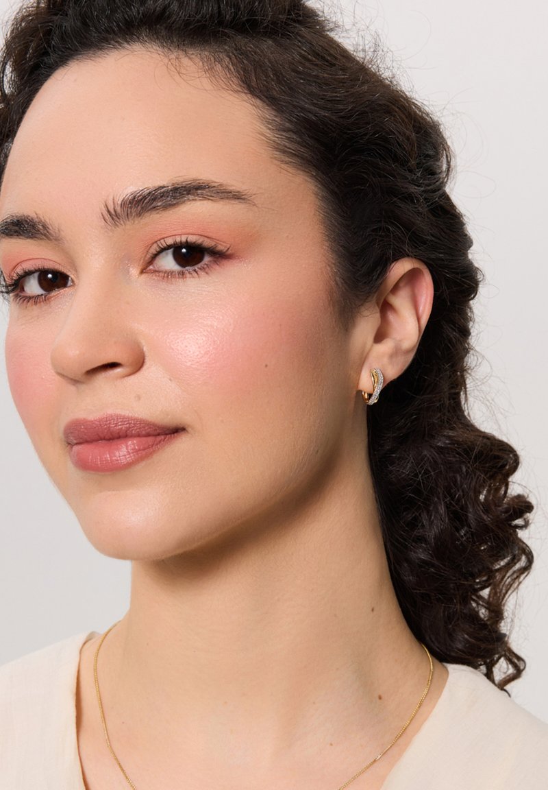 Złote kolczyki w kształcie obrączek o zaokrąglonym designie, z małym zdobieniem. Gładka tekstura; noszone w przekutym uchu. Ton skóry wydaje się naturalny.