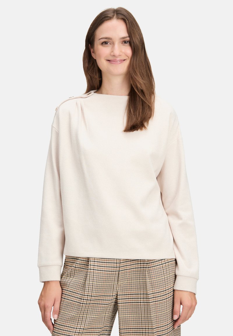 Jasnobeżowy sweter z okrągłym dekoltem, długimi rękawami i guzikami na ramionach, zestawiony z kraciastymi spodniami w wzór kratki.