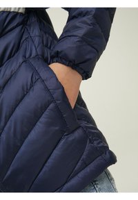 Marinblå puffjacka med en quiltad design, elastiska manschetter och en sidoficka. Materialet verkar blankt med en slät textur.