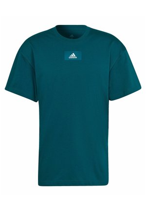 Homme portant un t-shirt Adidas teal avec logo sur la poitrine et un short beige, debout avec les bras détendus le long du corps.
