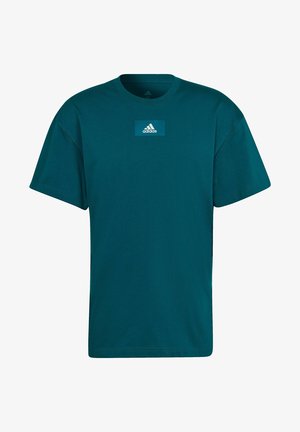 Homme portant un t-shirt Adidas teal avec logo sur la poitrine et un short beige, debout avec les bras détendus le long du corps.