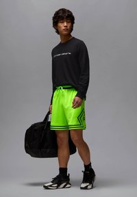 Chemise noire à manches longues associée à un short néon vert vif avec des accents noirs, tenant un sac de sport noir, portant des baskets noires et blanches.