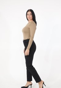 Femme aux cheveux longs et foncés portant un haut beige à manches longues, un pantalon noir taille haute et des chaussures à talons hauts noires, debout de profil.
