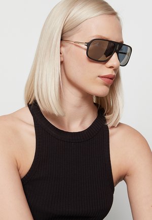 Donna bionda con capelli lisci indossa grandi occhiali da sole neri e un top nero senza maniche a coste, guardando leggermente a destra.