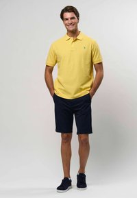 Žluté bavlněné polo tričko s límcem a zeleným logem, v kombinaci s námořnickými šortkami a námořnickými teniskami. Hladké textury bez vzorů.