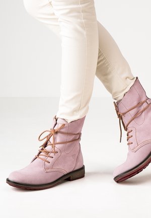Jasnoróżowe zamszowe botki do kostki z brązowymi sznurowadłami, noszone z kremowymi dopasowanymi spodniami, na białym tle podłogi.