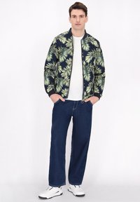 Giubbotto bomber con una base blu navy, caratterizzato da un motivo di foglie di palma verde chiaro. Abbinato a una t-shirt bianca e jeans blu ampi. Sneakers bianche.