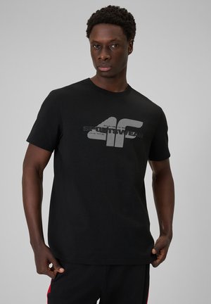 Mężczyzna w czarnej koszulce z krótkim rękawem z dużym białym i szarym logo "4F SPORTWEAR" na piersi na jednolitym szarym tle.