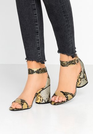 Füße, die offene Blockabsatz-Sandalen mit Schlangenprint und Knöchelriemen tragen, kombiniert mit schwarzen Jeans mit ausgefranstem Saum.