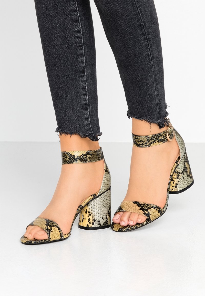 Pieds portant des sandales à talons blocs ouvertes à l'avant avec imprimé serpent et brides à la cheville, associées à un jean noir à ourlet effiloché.