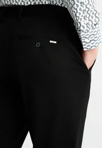 Schwarze Hose mit einer geknöpften Gesäßtasche, aus glattem Stoff gefertigt. Ein kleines Etikett mit der Marke ist auf der Tasche sichtbar.