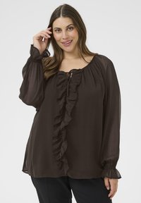 Mujer con una blusa de manga larga marrón oscuro con volantes en el frente y en los puños, sonriendo con una mano cerca de su cabello.