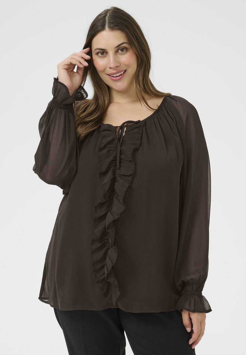 Mujer con una blusa de manga larga marrón oscuro con volantes en el frente y en los puños, sonriendo con una mano cerca de su cabello.