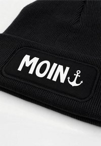 Black knit beanie with a rectangular patch featuring the word "MOIN" in bold white letters and an anchor icon, textured fabric detail.