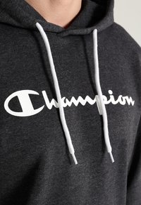Mörkgrå hoodie med vita dragsnören och "Champion"-logotyp i vit kursiv text över bröstet, bärs av en person.
