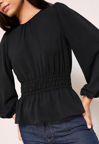 Blusa nera con maniche lunghe a palloncino, vita arricciata e orlo peplo. Il tessuto testurizzato aggiunge un tocco di dettaglio. Abbinata a jeans blu.