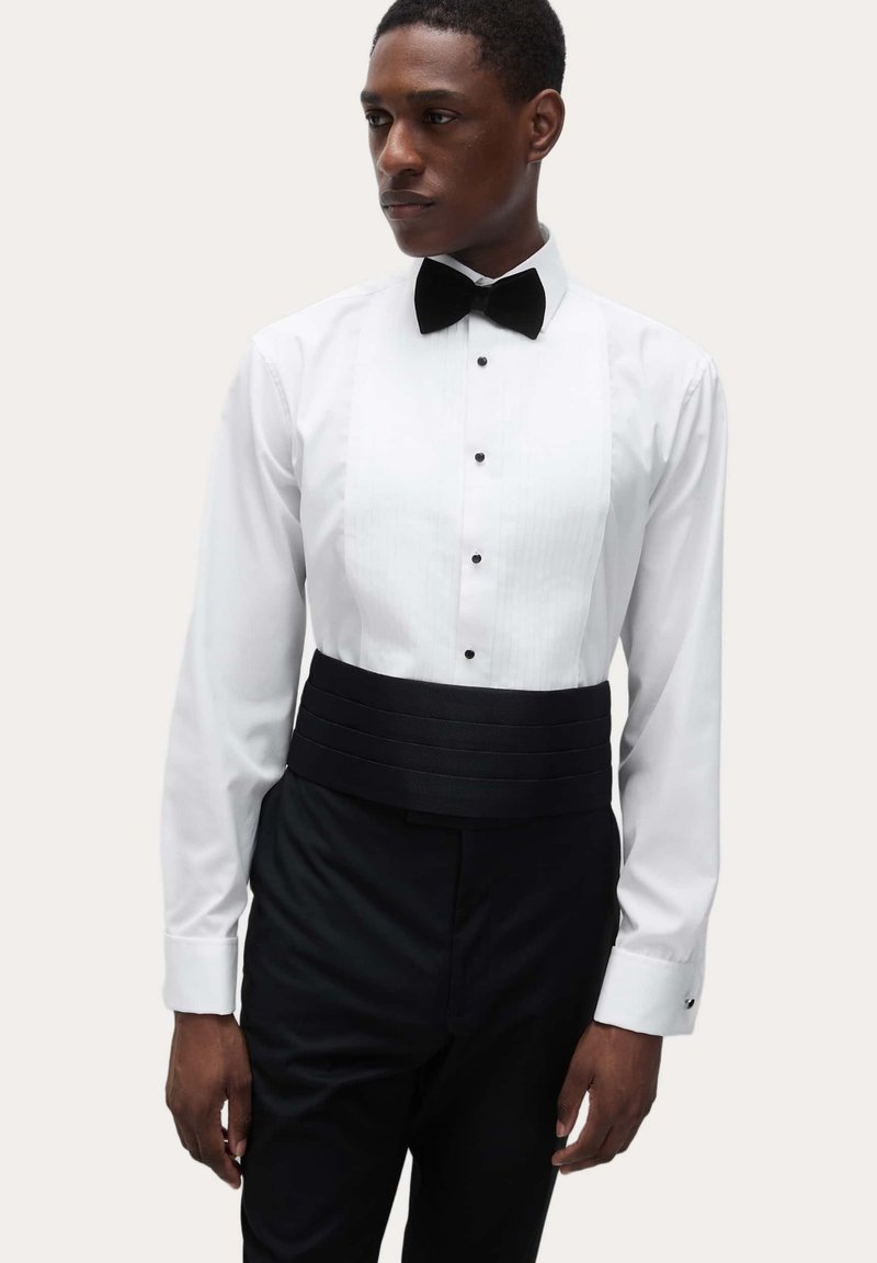 Hvid smoking-skjorte med sort butterfly, der har et plisseret frontpanel og sort taljebånd. Lange ærmer med manchet-detaljer, skræddersyet pasform.