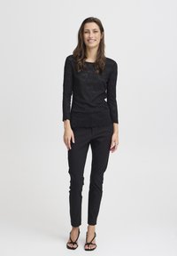 Top lungo in pizzo nero con maniche lunghe e orlo a scalloped, abbinato a pantaloni neri slim-fit e sandali neri con cinturini. Tessuto liscio e testurizzato.