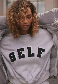 Pelēks sporta krekls ar melniem burtiem "SELF"; tam ir ribota apkakle un piedurknes. Gaiši pelēks jaka daļēji pārklāta uz pleciem.