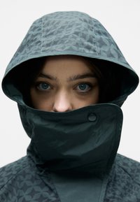 Tume roheline jakk mustrit paisutava kapuutsiga. Omab kõrget kaelanõlva ja ees kinnitust nööbiga. Sile, veekindel kangas.